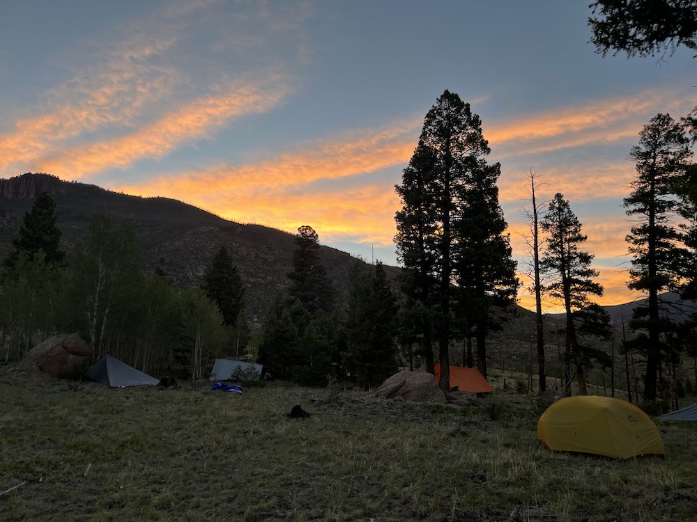 The sun sets over a mountain range with a cotton candy-colored sky. A row of tents and trees sit in the darkened foreground.